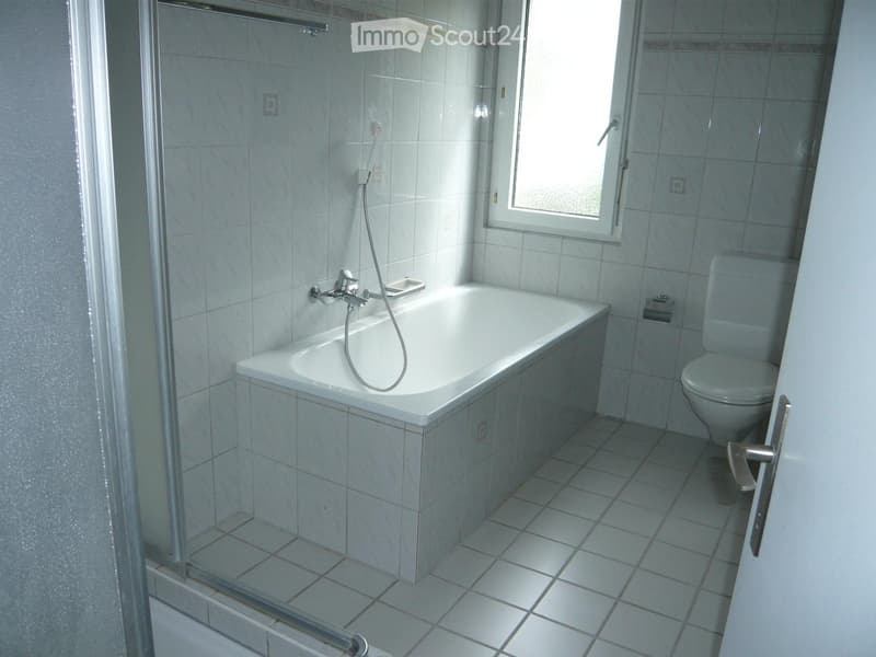 Modern bathroom with a white bathtub, toilet, and glass-enclosed shower. The room features tiled walls and floor, and a window providing natural light.