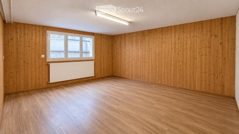 Empty room with wooden walls and floor, large window, radiator, and ceiling light fixture.