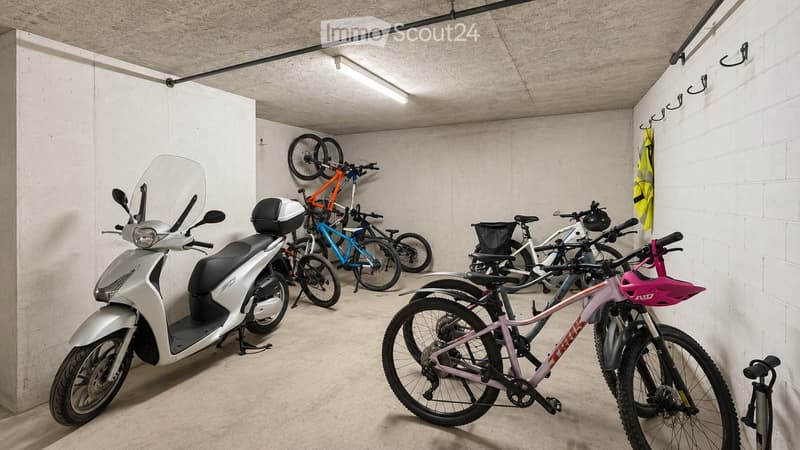 A modern, clean, and organized parking area with a scooter, several bicycles, and helmets neatly stored. The space features concrete walls and a concrete floor, with a few hooks and a safety vest visible.