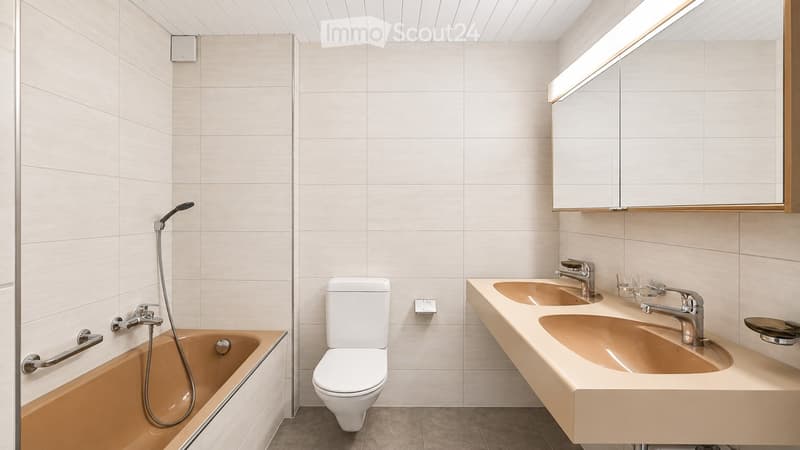 Modern bathroom with a bathtub, toilet, double sink, and a large mirror with built-in lighting. The walls are tiled in light beige, and the floor is tiled in a neutral tone.