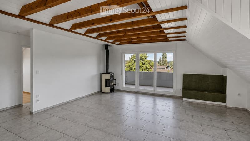 Empty room with white walls, exposed wooden beams, tiled floor, and a fireplace. Large sliding glass doors lead to a balcony with a view of greenery.