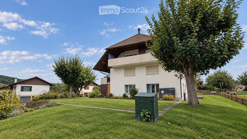 Two-story house with a balcony, surrounded by a well-maintained lawn and trees. The house features a tiled roof and multiple windows.