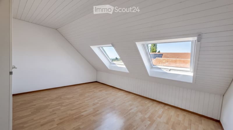 Empty attic room with wooden flooring, white paneled walls, and skylight windows offering a view of the roof and surrounding area.
