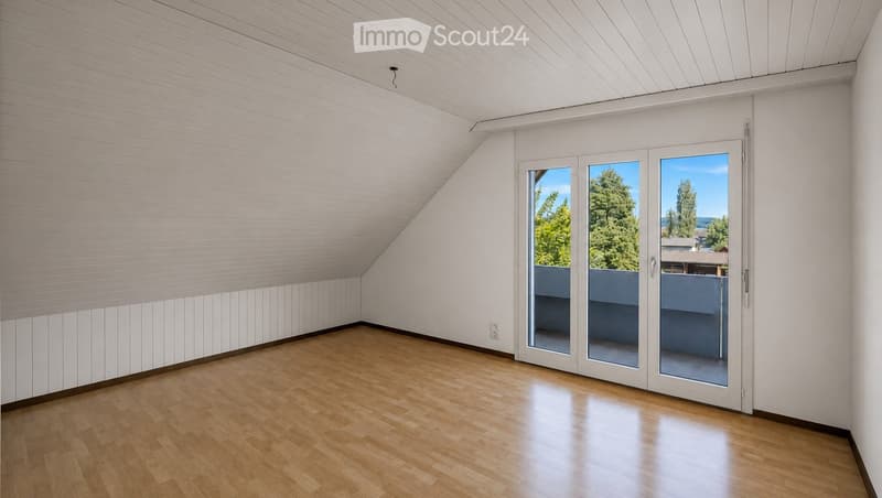 Empty room with wooden flooring, white paneled walls, and large sliding glass doors leading to a balcony with a view of trees and a house.