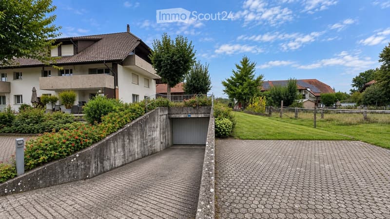 Two-story house with a tiled roof, white exterior, and a balcony on the upper floor. The house features a driveway, a garage entrance, and is surrounded by greenery and a well-maintained lawn.