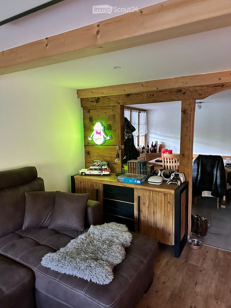 A cozy living room with a brown leather sofa, a wooden coffee table, a gaming console, and a furry blanket on the sofa. The room features exposed wooden beams and a modern aesthetic with a mix of furniture and decor.