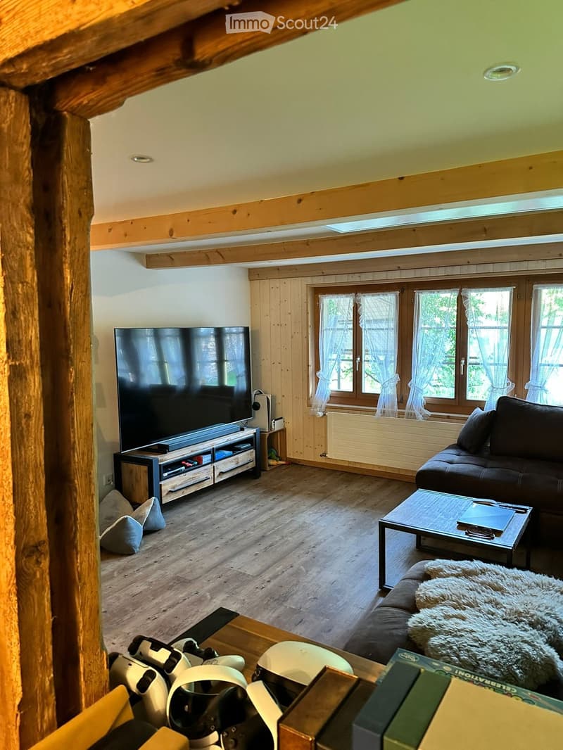 A cozy living room with wooden walls and floor, featuring a large television on a stand, a dark brown sofa with throw pillows, a coffee table, and gaming equipment on a side table.