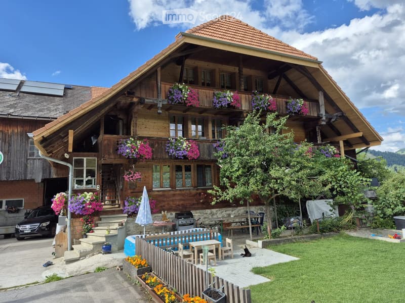 Traditional wooden house with a sloped roof, multiple windows, and flower boxes on the balcony. The house has a well-maintained garden with a patio, outdoor furniture, and a small swimming pool.