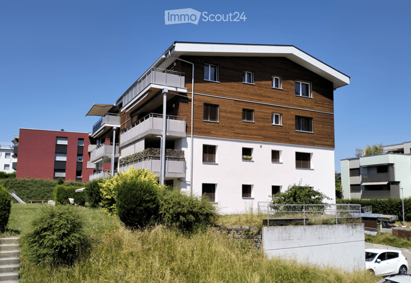 Modern multi-story residential building with balconies, wooden facade, and surrounding greenery.