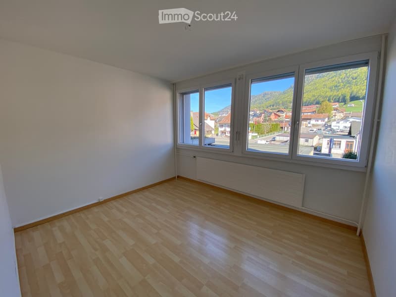 Empty room with wooden flooring, white walls, large windows showing a scenic view of mountains and town, radiator under windows.
