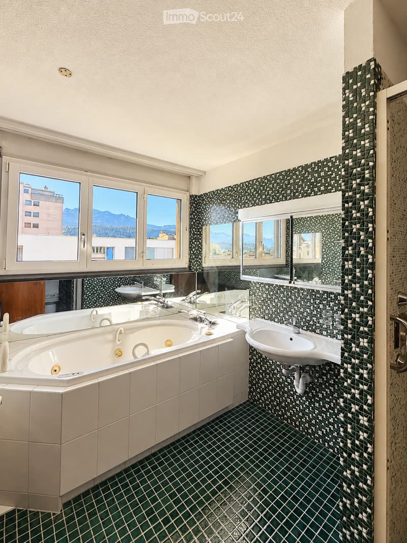 Modern bathroom with a large bathtub, double sink, and large windows offering a scenic mountain view. The walls are tiled in a black and green mosaic pattern, and the floor features green tiles with a diamond pattern.