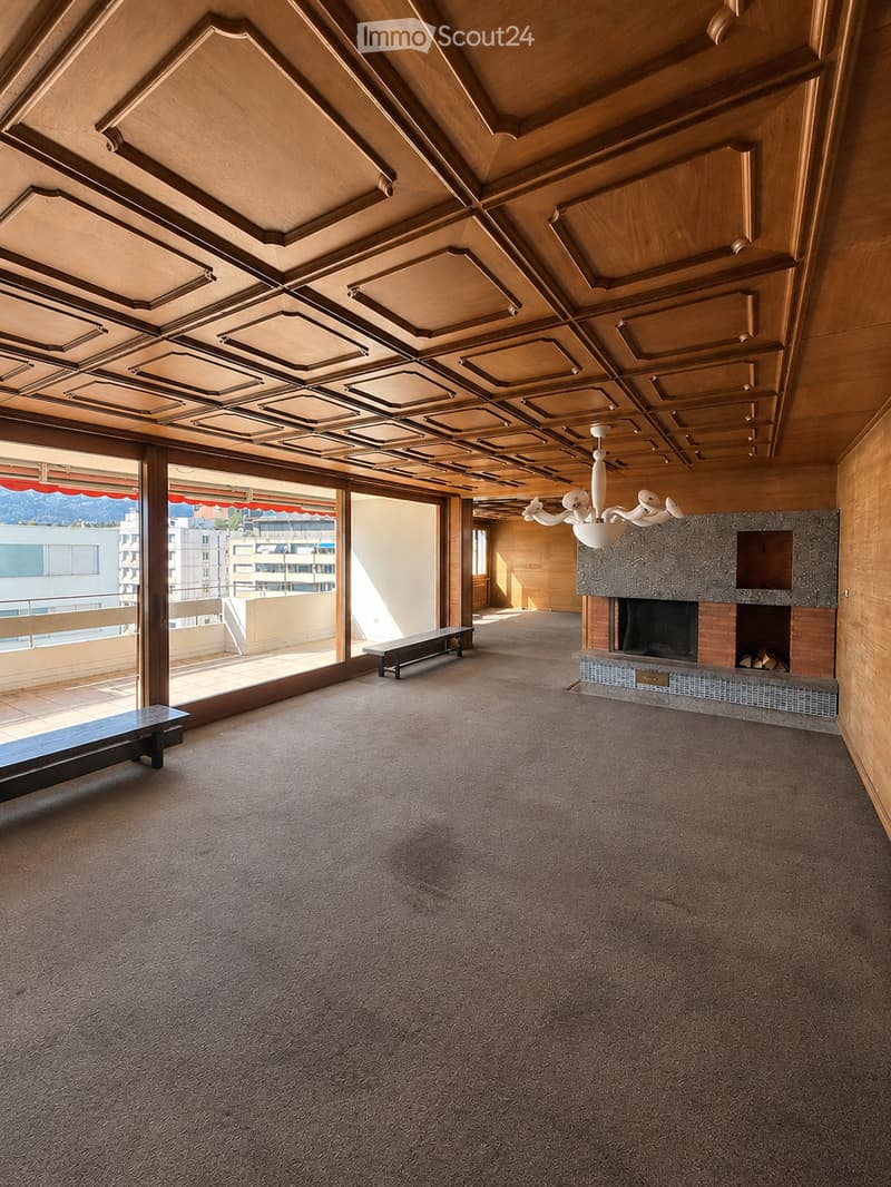 Spacious living room with a decorative wooden ceiling, large windows offering a view, a fireplace, and two benches.