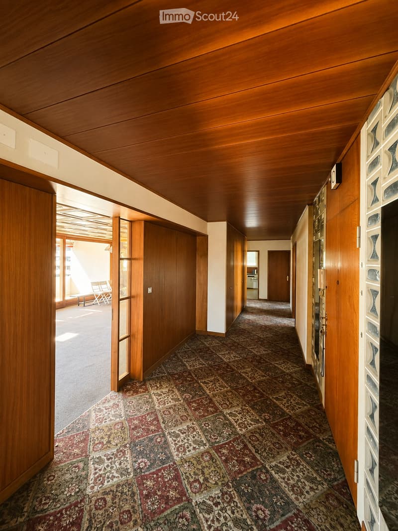 A long, narrow hallway with wooden paneling on the walls and ceiling, patterned carpet, and several doors leading to other rooms. Natural light streams in from an open doorway at the end.