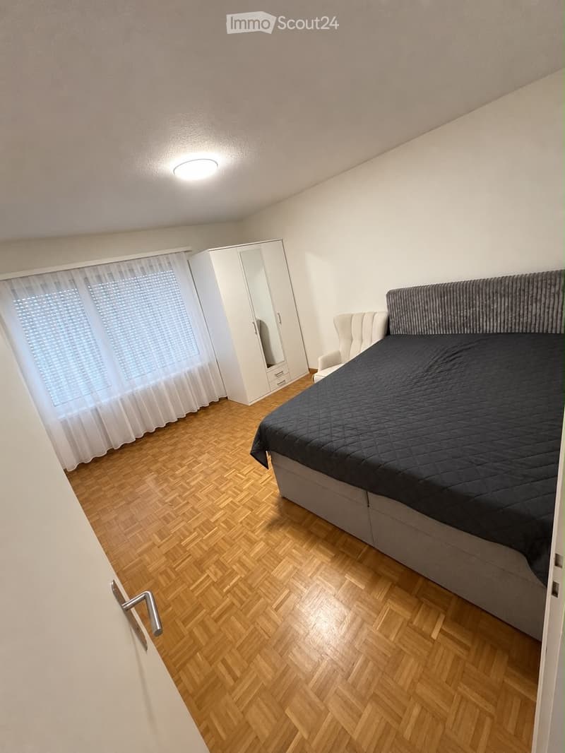 Modern bedroom with wooden herringbone flooring, a large bed with dark bedding, a white wardrobe with mirrored doors, and a white armchair near the window.