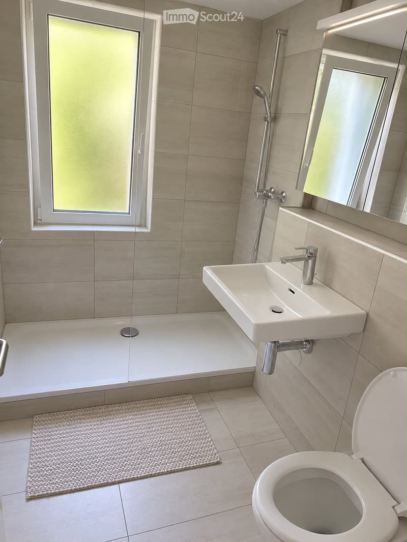 Modern bathroom with a bathtub, sink, toilet, and showerhead. Features a large window with frosted glass, beige tiles, and a small textured rug.