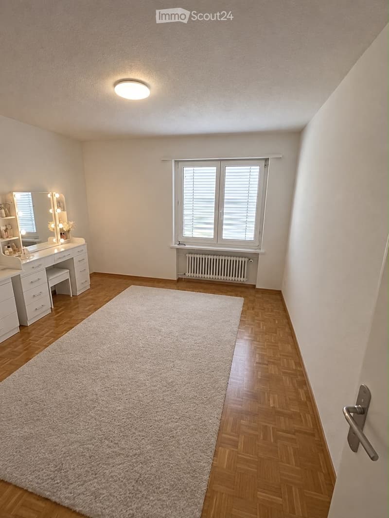 A well-lit room with wooden flooring, a large white shag rug, a vanity with a mirror, and a radiator under a window with blinds.