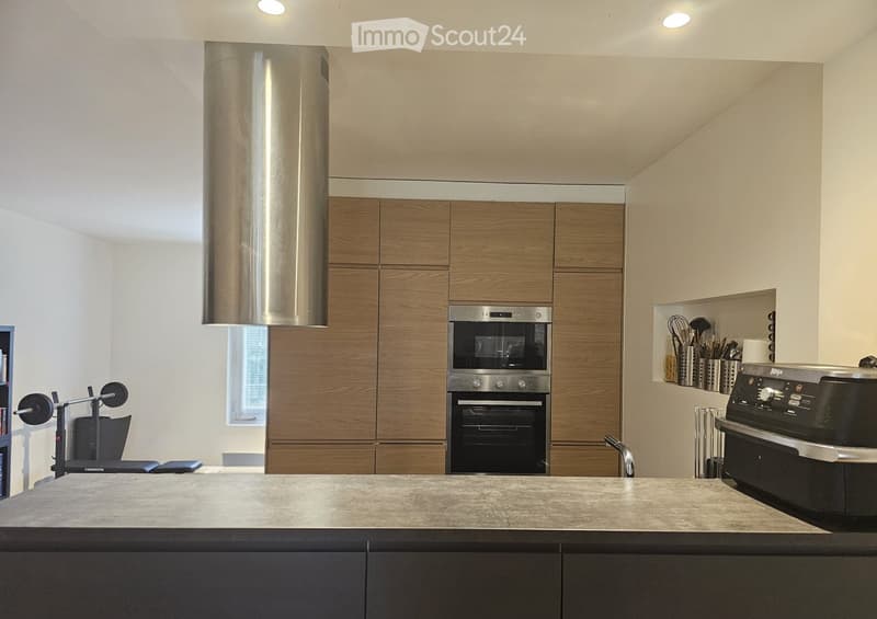 Modern kitchen with wooden cabinets, stainless steel appliances, a large island, and a built-in oven with a range hood. Exercise bike is visible in the background.