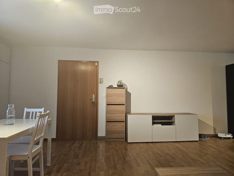 Dining room with a wooden dining table, chairs, a cabinet, and a white cupboard. The room has light-colored walls and wooden flooring.