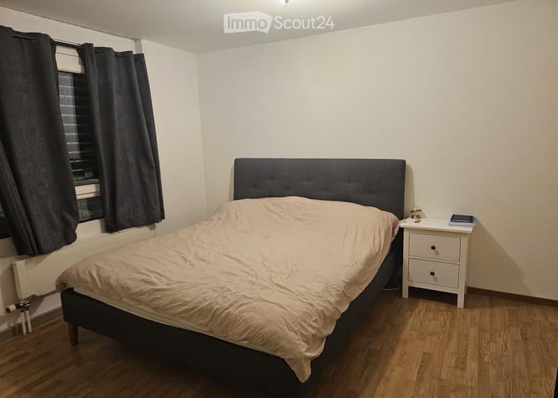 A simple bedroom with a single bed, a nightstand, and dark curtains on the window. The bed has a beige duvet and a dark headboard.