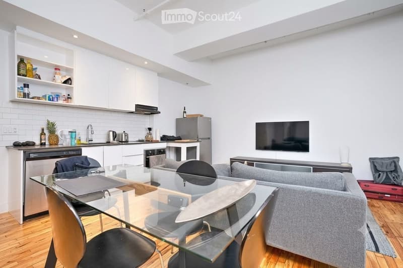 Open-plan living and dining area with a grey sofa, glass dining table, and modern kitchen featuring stainless steel appliances, white cabinets, and a countertop with various items.