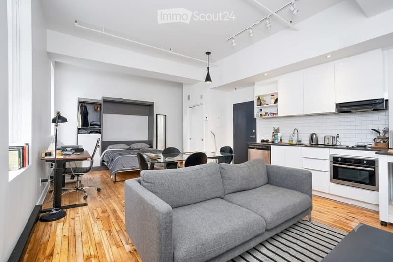 Modern, open-concept studio apartment with a grey sofa, dining table, bed, and a compact kitchen area featuring white cabinets and stainless steel appliances.