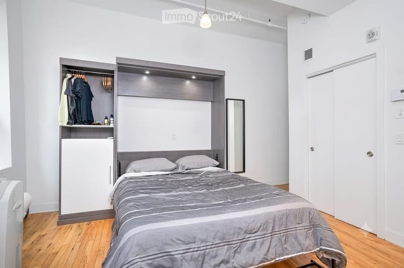 Modern bedroom with a grey duvet-covered bed, built-in wardrobe with hanging clothes, a small refrigerator, and a mirror on the wall.