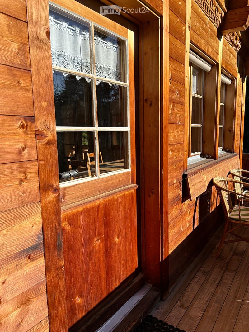 Wooden house exterior, large window with wooden frame, balcony with wooden chair and table, wooden flooring