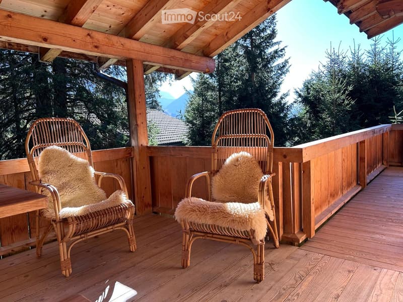 covered balcony with wooden deck, rattan chairs, small table, wooden railing, trees, mountains in the distance
