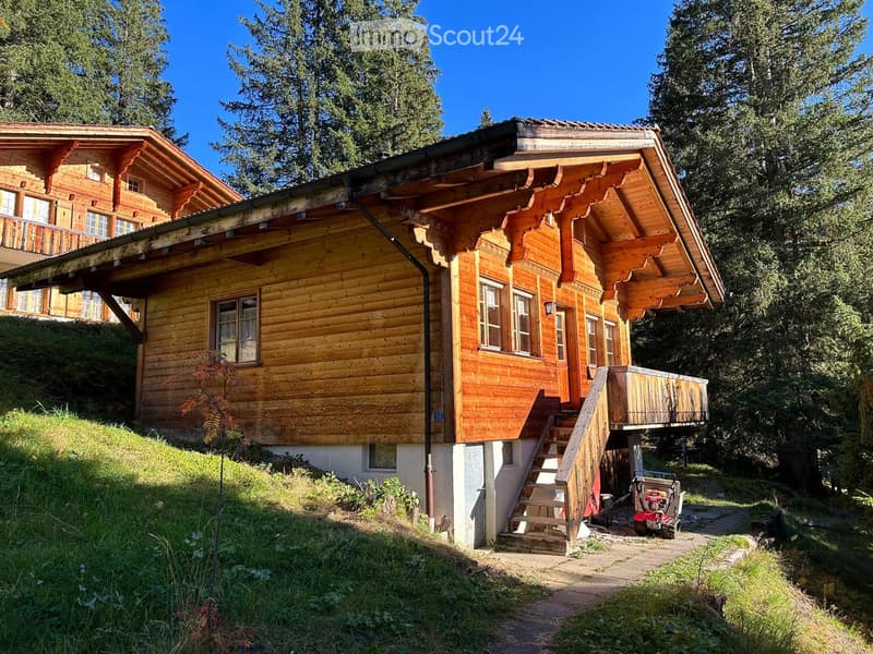 wooden chalet, tiled roof, wooden balcony, stairs leading to entrance, glass windows, surrounded by trees
