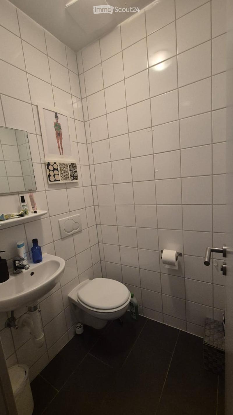 A compact bathroom with white tiled walls and dark tiled floor, featuring a toilet, sink, mirror, and a small shelf with toiletries.