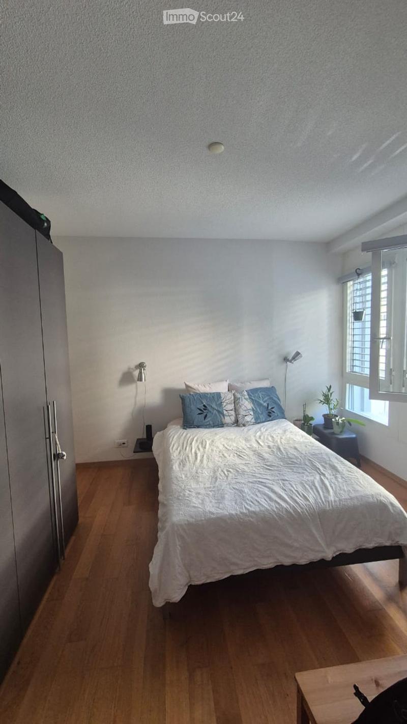 A small, well-lit bedroom with a wooden floor, a single bed with white bedding and blue pillows, a nightstand with a plant, and wall-mounted lamps.