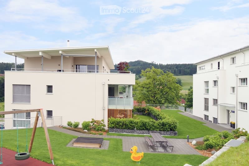 Modern two-story residential building with a balcony, surrounded by a well-maintained lawn, playground equipment, and a picnic table.