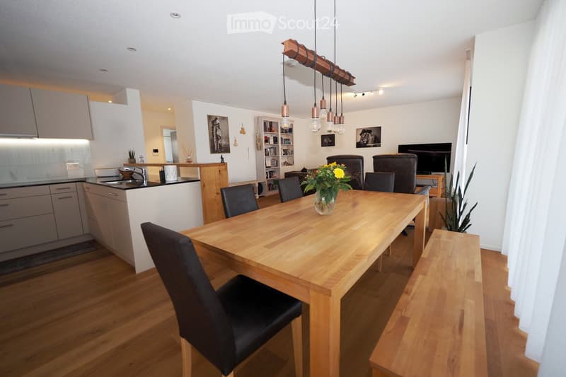 Open-plan living and dining area with a wooden dining table, black chairs, a modern kitchen counter, and a television area with a sofa.