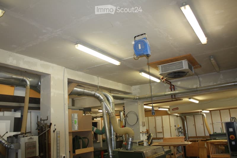 The image shows a workshop or industrial space with exposed pipes, a large air conditioning unit, fluorescent lighting, and various tools and equipment, including a table and cabinets.