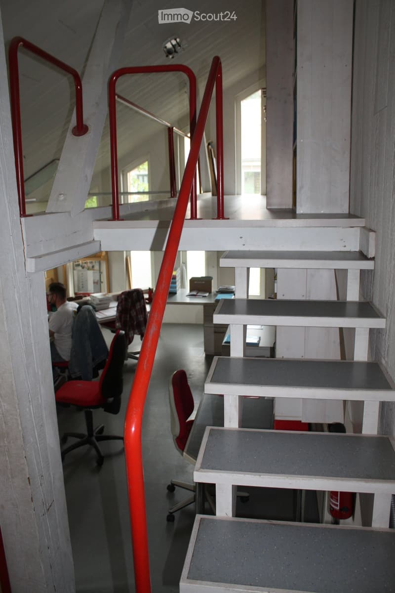A modern, open staircase with red railings leads to an upper level. The lower level features a workspace with a desk, chairs, and office supplies.