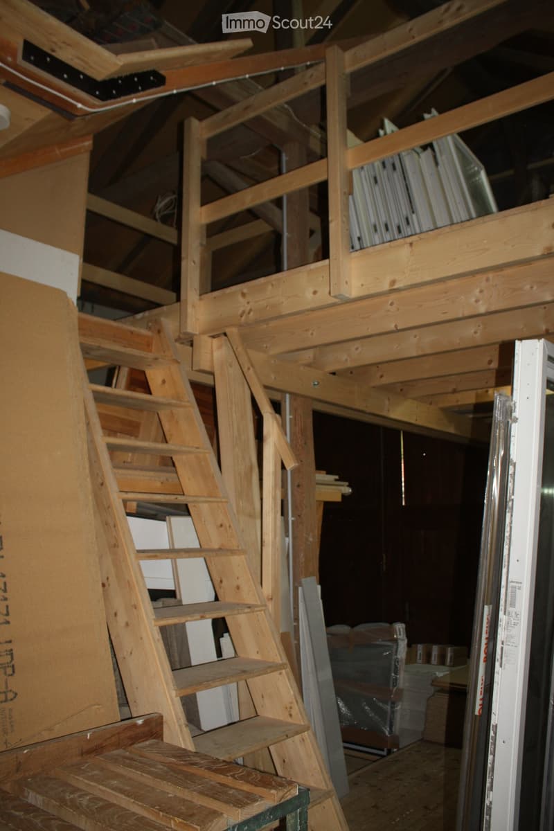 An unfinished attic space with exposed wooden beams, a wooden staircase, and various construction materials and tools visible.