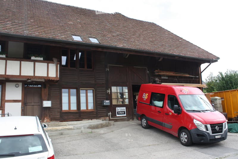 Traditional wooden building with a steeply pitched roof, large windows, and a balcony. A red van is parked in front, and there is a sign on the building.
