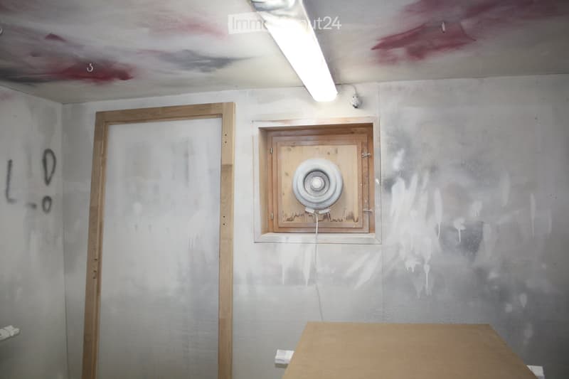 Empty room with unfinished walls and ceiling, featuring a wooden door frame, a ventilation unit, and a simple wooden table.