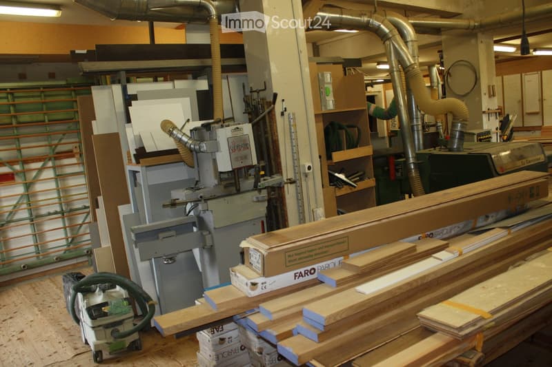 A workshop filled with woodworking tools and materials, including a large woodworking machine, stacks of wood planks, a dust collector, and storage shelves.