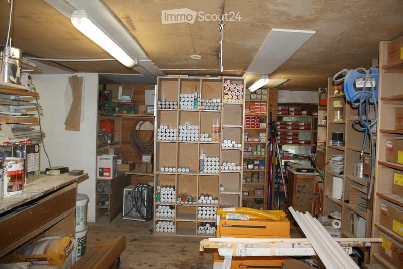 A cluttered workshop or storage room with wooden shelves filled with tools, paint cans, and boxes. The space includes a workbench, various storage cabinets, and a variety of construction materials.