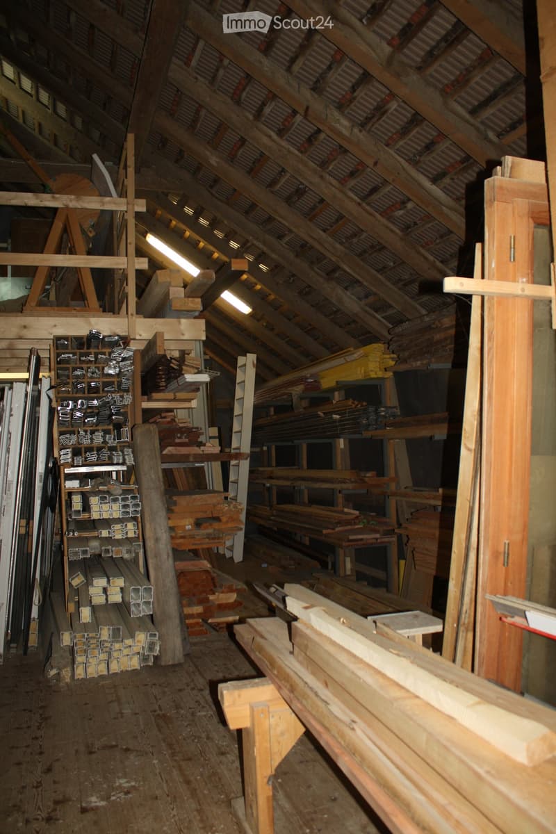 An attic space with exposed wooden beams and metal roofing, containing stacks of wooden planks, metal bars, and construction materials on shelves and workbenches.