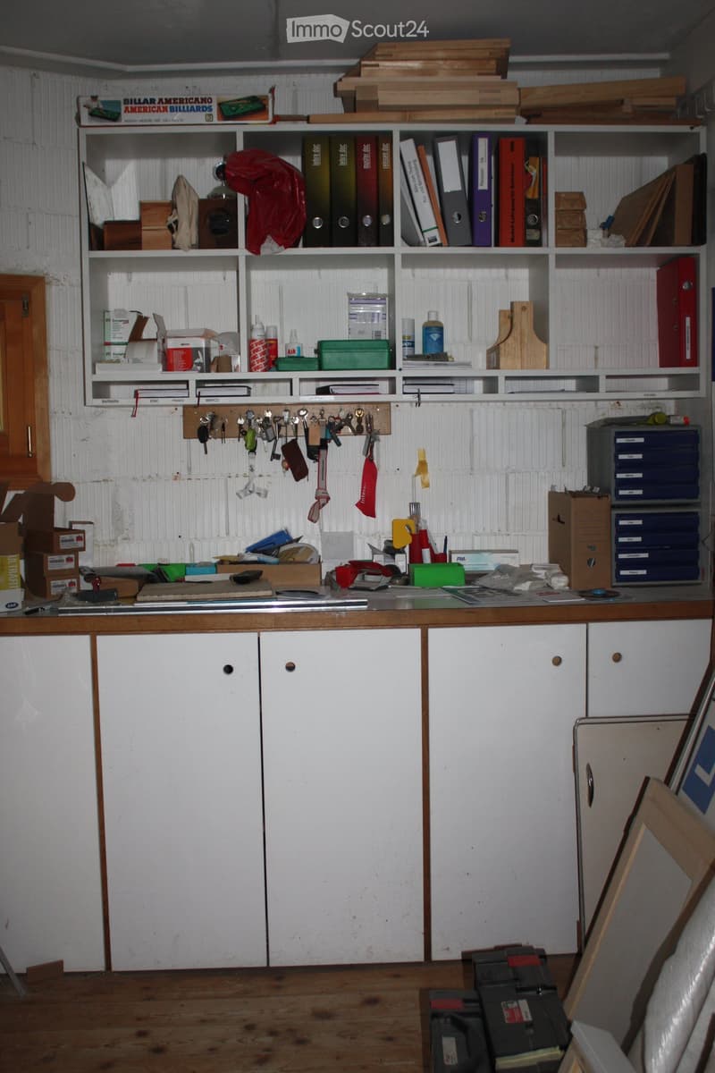 A cluttered room with a wooden table, white cabinets, shelves filled with books, folders, and various items, and a key rack on the wall.
