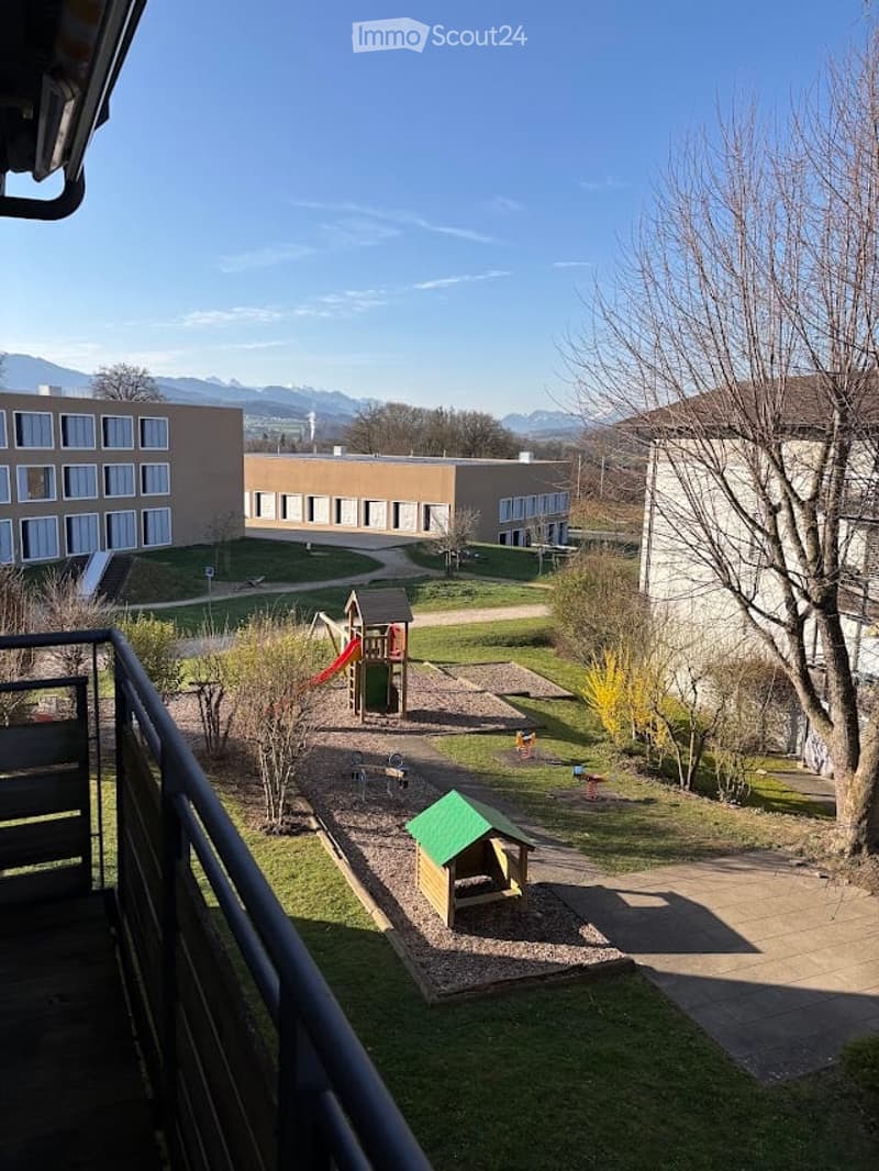 View from a balcony overlooking a playground area with slides, swings, and a small doghouse, surrounded by grassy patches and pathways, with buildings and mountains in the background.