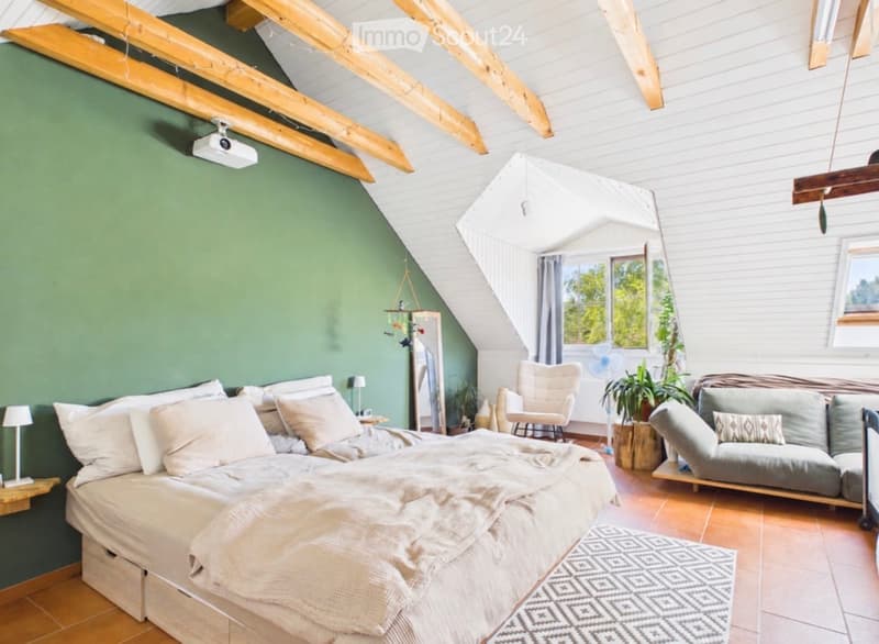 Cozy attic bedroom with exposed wooden beams, green accent wall, large bed, sofa, armchair, small table, and hanging plants.