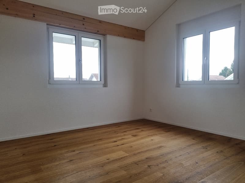 Empty room with wooden flooring, white walls, and two windows offering a view of rooftops.