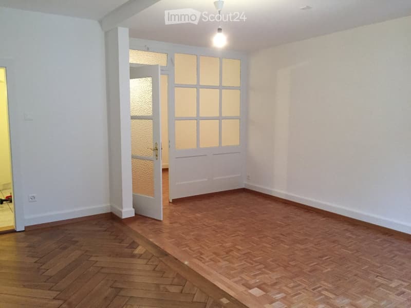 Empty room with wooden flooring, white walls, and a hanging light fixture. There is an open doorway leading to another room with a visible orange floor.