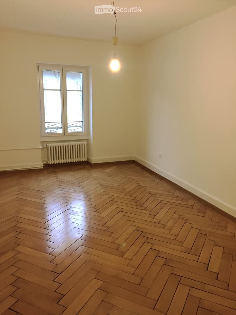 Empty room with wooden herringbone flooring, white walls, a single hanging light fixture, a radiator, and a window with double panes.
