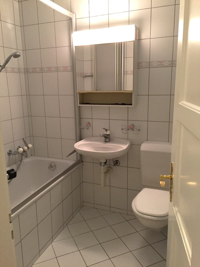 Clean, modern bathroom with white tiles, bathtub, toilet, sink, mirror, and showerhead.