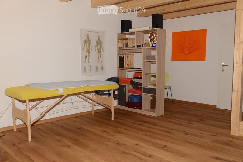 A well-lit room with wooden flooring, a massage table covered in a yellow and blue sheet, a wooden shelf holding various items, a green chair, and posters on the wall.