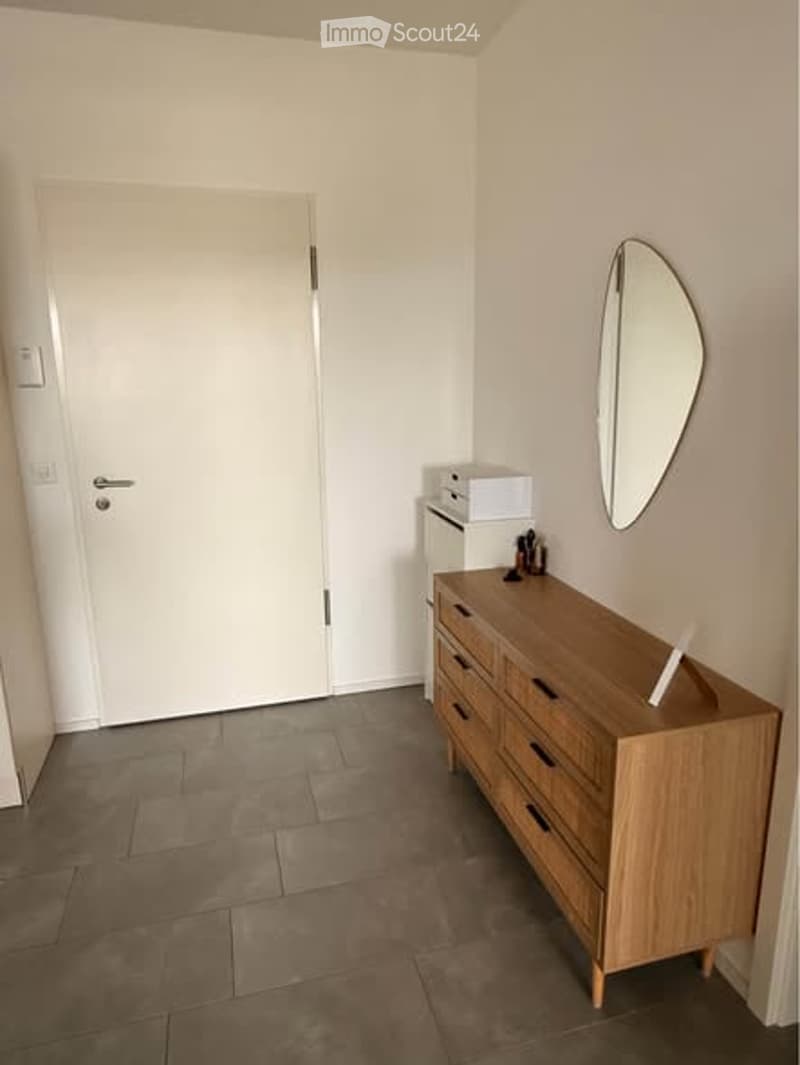 A small, neatly furnished room with a wooden dresser, a round mirror, and a small cabinet. The room has a tiled floor and white walls.